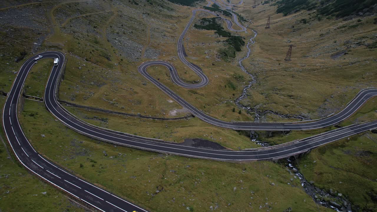 A graceful left-to-right sweep reveals the winding curves of Transfagarasan road, weaving through lush green mountains in Romania under a moody summer sky.