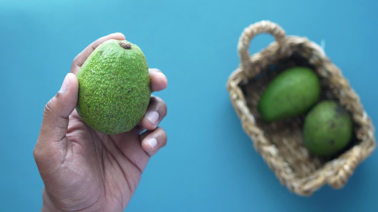 mano sosteniendo un aguacate, con otros aguacates en una canasta
