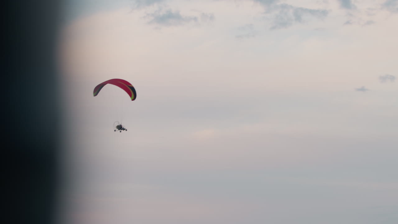 Silhouetted view of people flying powered paraglider under pastel evening sky, red wing arched above suspended trike as they drift through soft fading light with scattered clouds