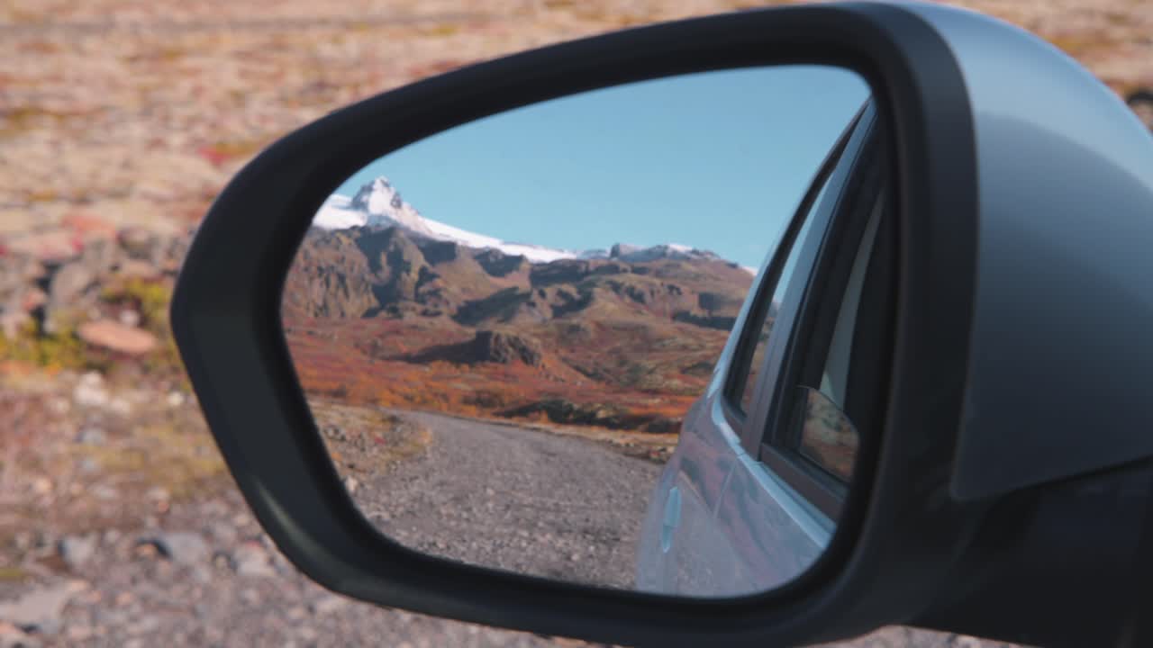 Car side-view mirror reflecting mountain panorama and road in Iceland