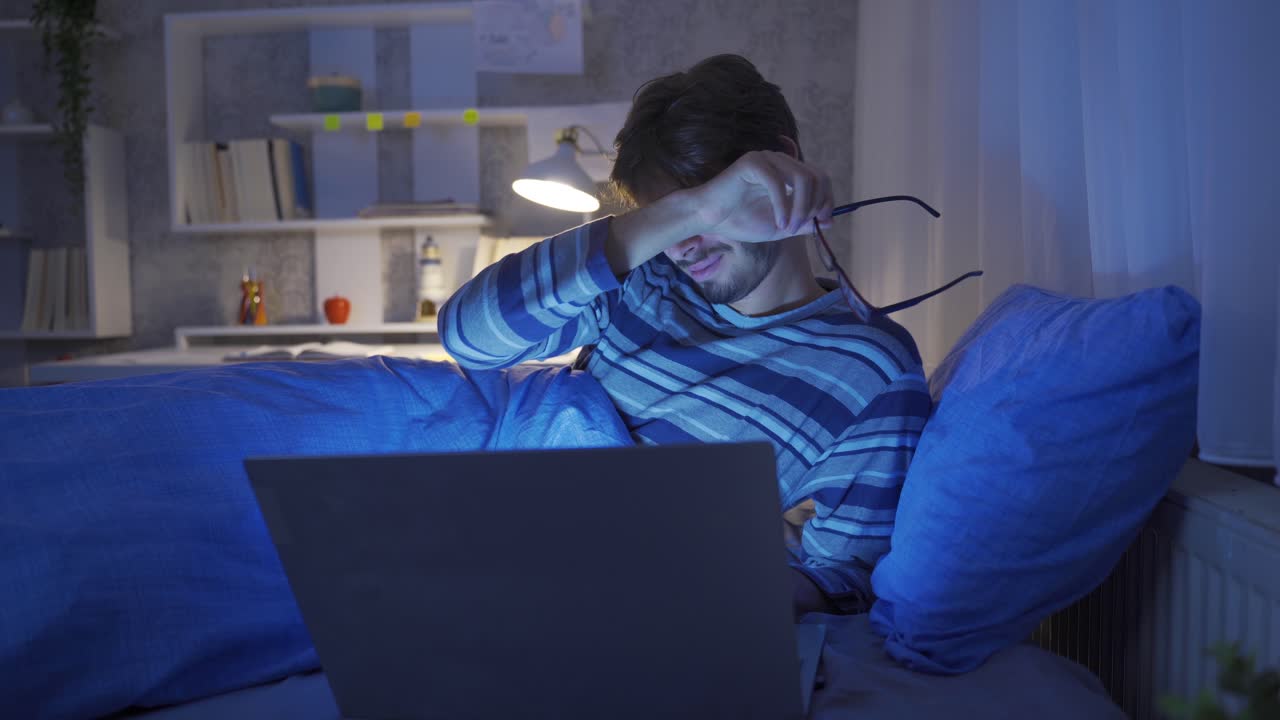 Young man working with laptop in bed. He is tired and sleepy.