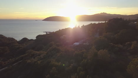 Tiro-Aéreo-De-Drones-Rastreando-Hacia-Atrás-Sobre-Una-Casa-Con-Energía-Solar-Y-árboles-Cubiertos-De-Luz-Solar-Dorada-Durante-La-Puesta-De-Sol-En-Elba,-Italia