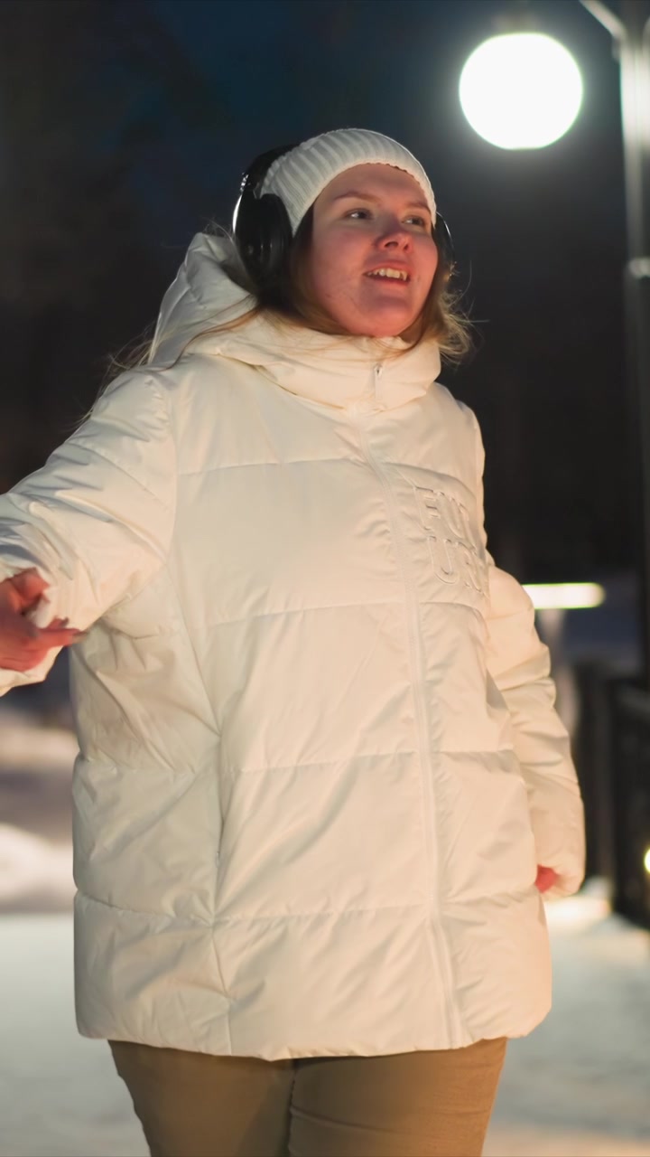 Young woman wearing white puffer jacket and black headphones dances outdoors at night with soft lighting illuminating her movements. Woman is seen from various angles as woman performs dance