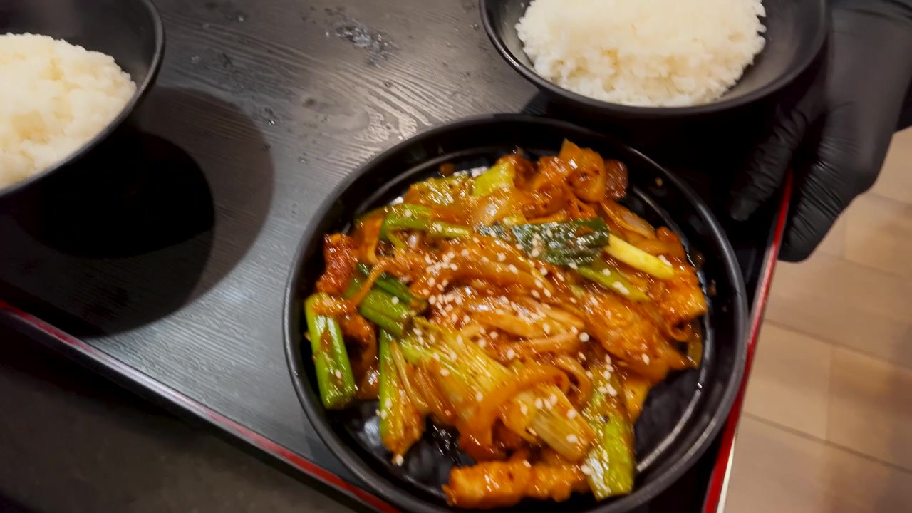 Bidikan dari atas seorang pelayan menata tumisan daging babi, kimchi, dan nasi di atas nampan.