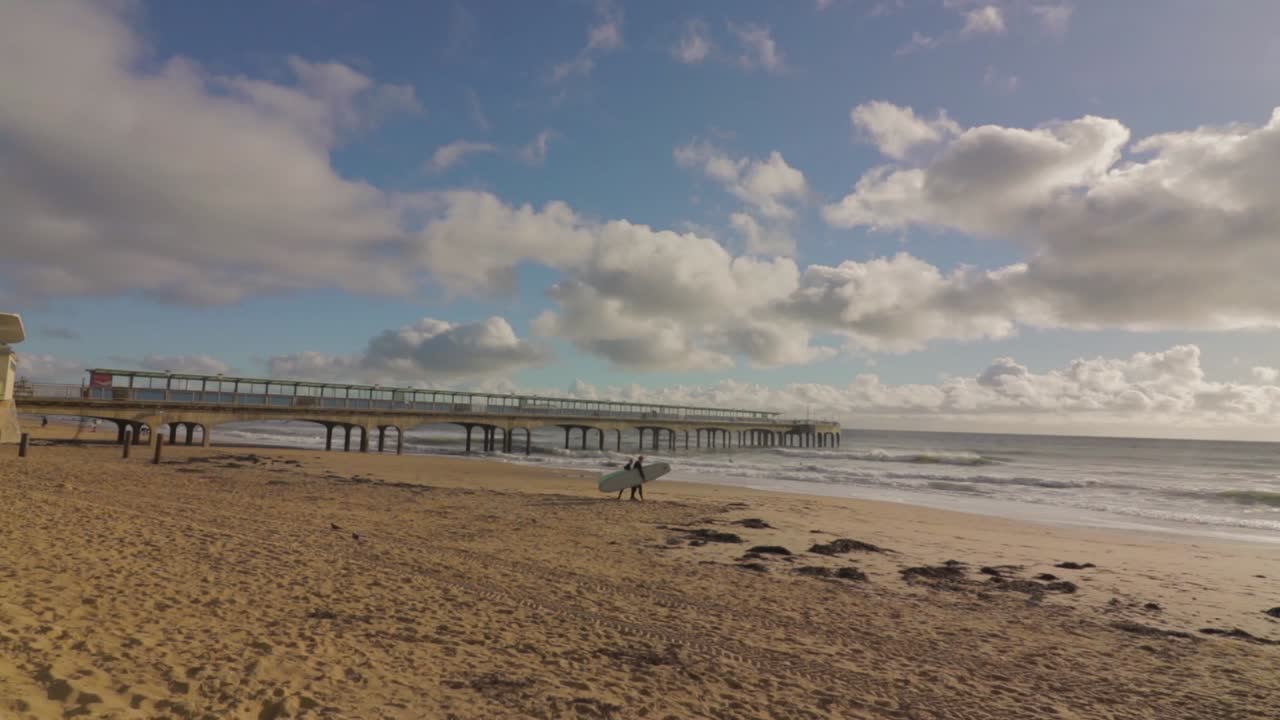 Two surfers with boards, walking on the beach towards group of surfers