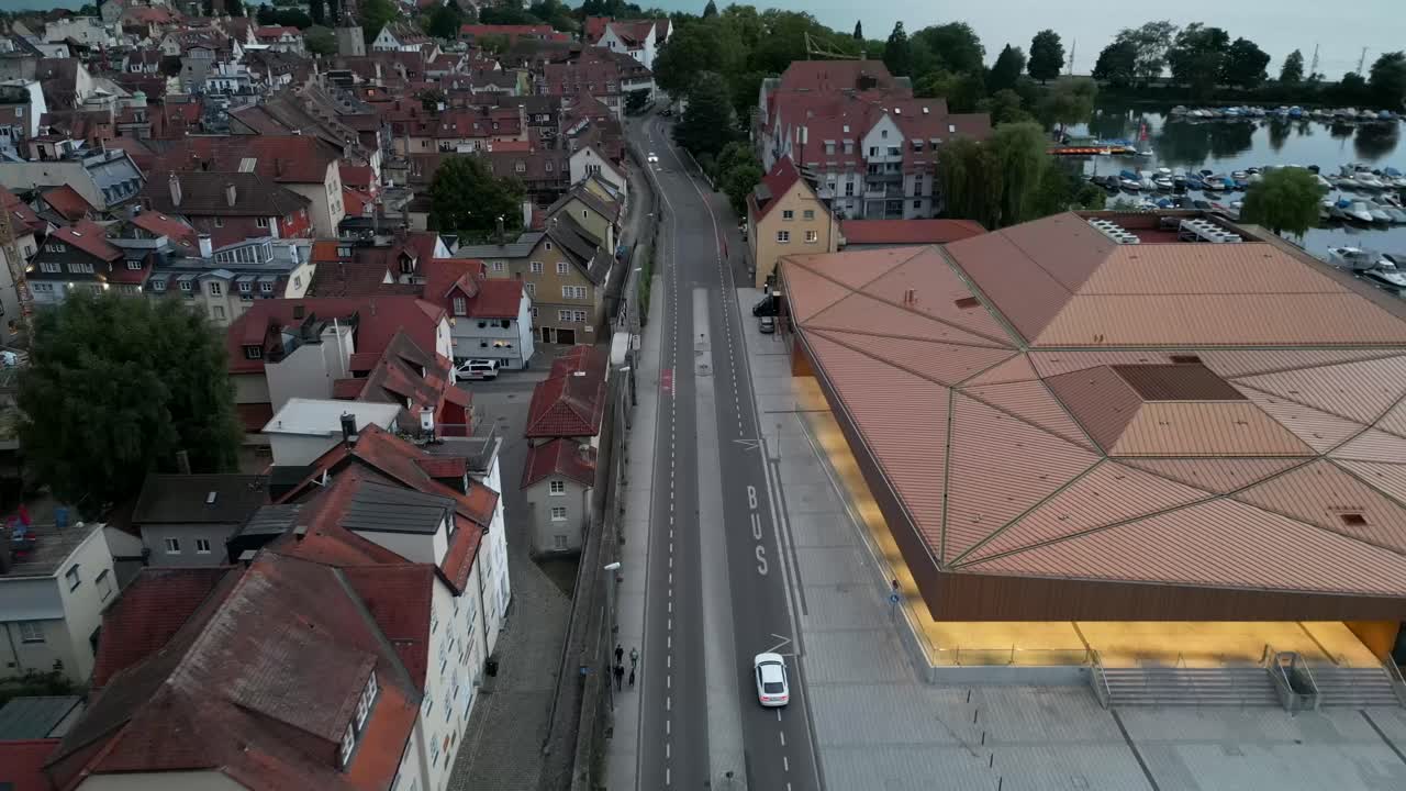 Drone footage spans a road with commercial and residential buildings on the left and a harbor filled with boats on the right, offering stunning views