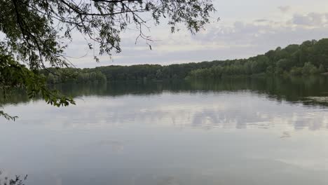 Pequeño-Lago-Cerca-De-Colonia-Con-Aguas-Tranquilas-Con-árboles-Alrededor-Y-En-La-Orilla-Bajo-Un-Cielo-Nublado-Con-Nubes