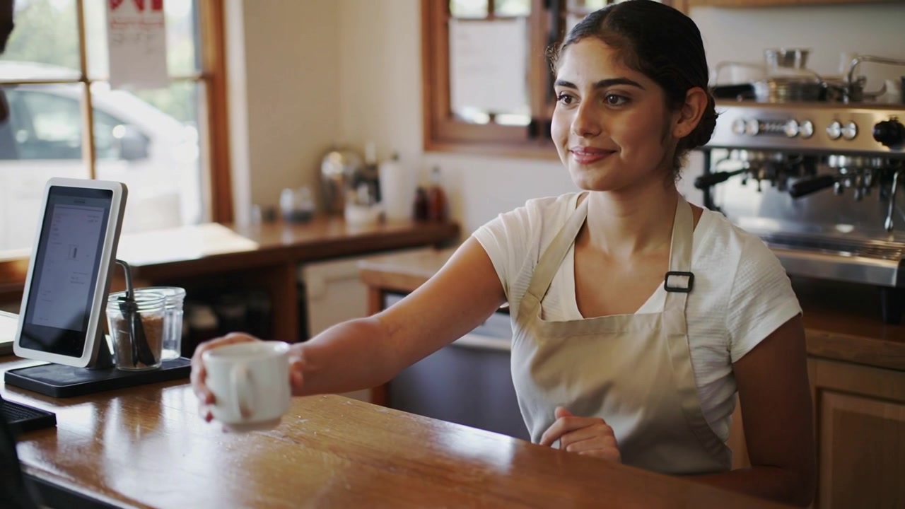 Sliding barista in white shirt beige apron moving white cup across wood counter, customer reaching