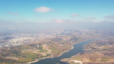 During-daylight,-a-view-of-Istanbul-city-from-the-airplane-window,-showcasing-the-Sazlidere-Dam-meandering-amidst-buildings-and-vacant-lots,-with-a-partly-cloudy-sky