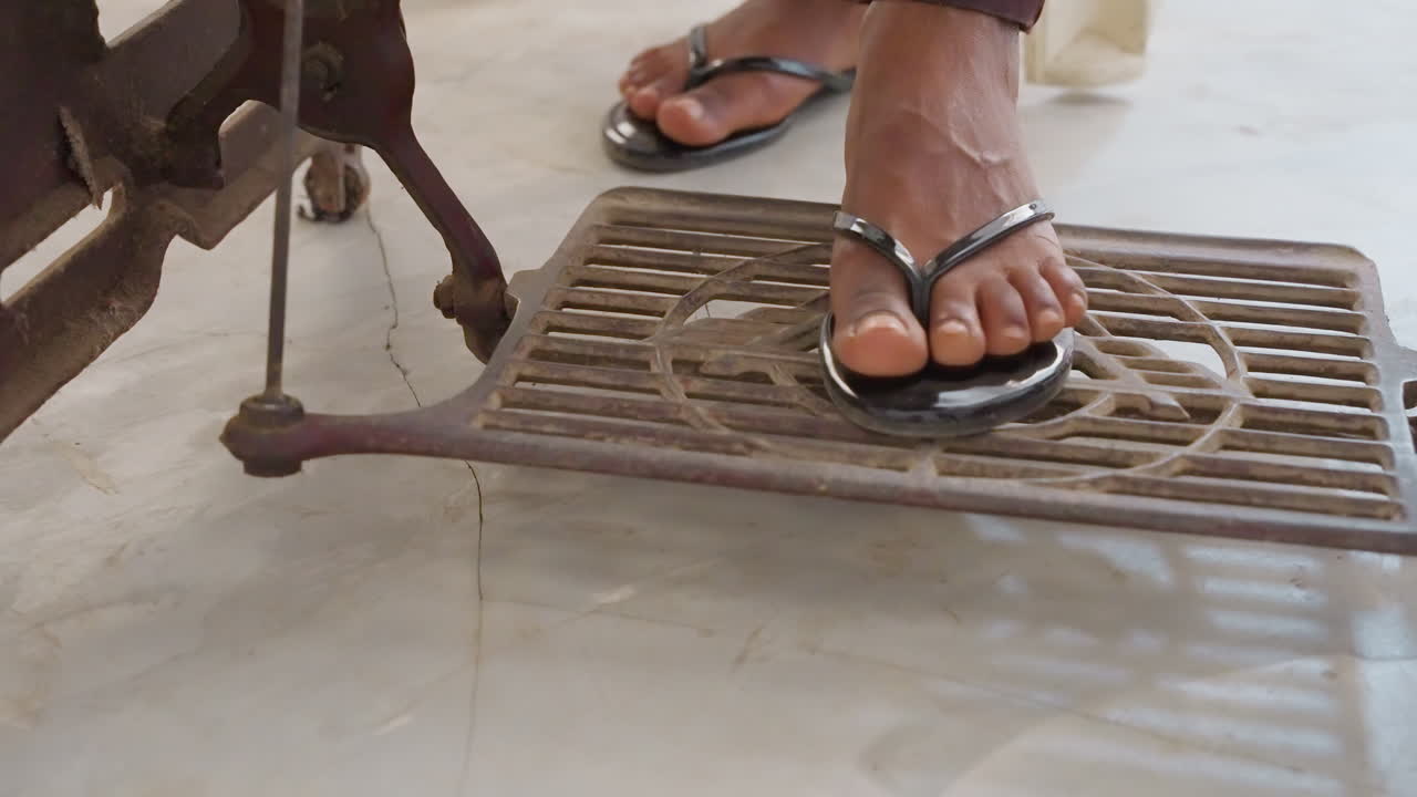Primo piano: sandalo su pedale a bilanciere di un'antica macchina da cucire nera, sul pavimento di casa illuminato dal sole, piede che preme con movimento ritmico, punta che afferra il cinturino del sandalo, motivo in ghisa, tranquillo lavoro domestico, capo fatto a mano.