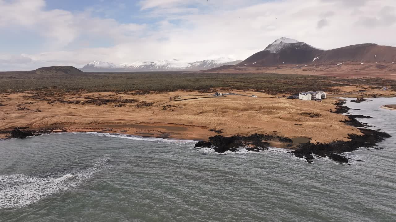 Drone view of Icelandic coast near Borgarfjörður with ocean waves, rocky shore and snow mountains in background.