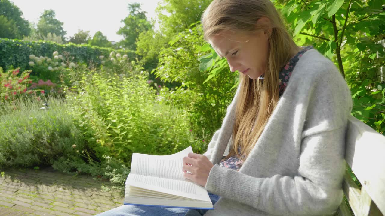 Slow motion medium shot, from the side, of an attractive young woman reading and studying in a lush green garden environment turning pages
