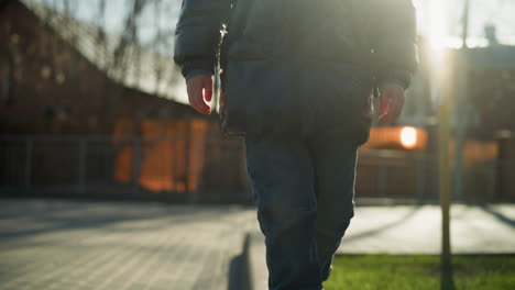 a close-up of a child carefully walking along a pavement, dressed in a shiny black jacket, with blurred trees and buildings in the background, illuminated by soft sunlight