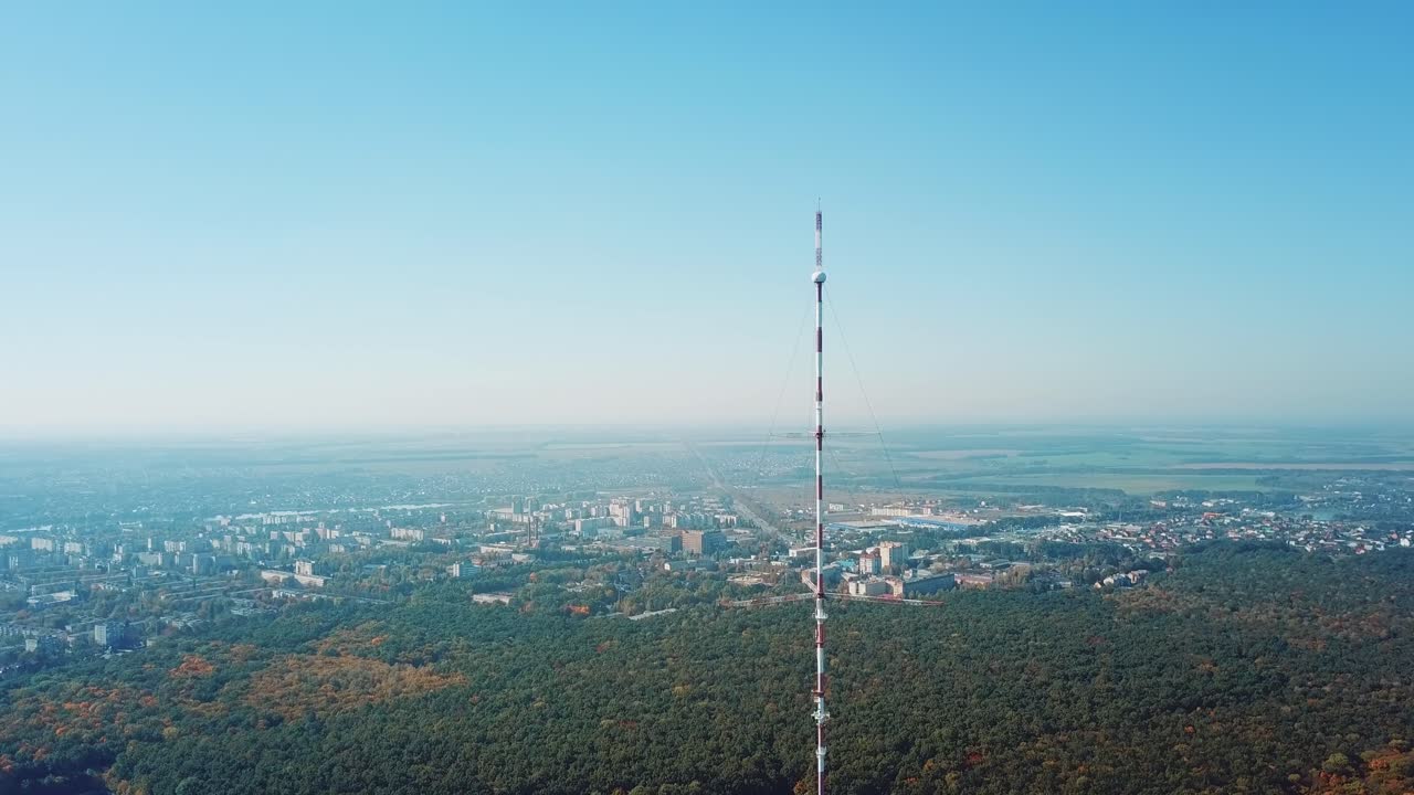top of the telecommunication tower in red and white colours on the background of the cityscape and blue sky. Camera motion up