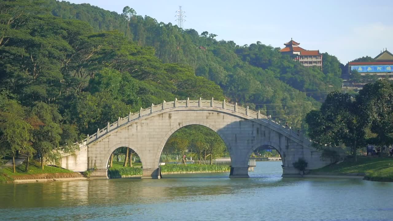 A peaceful bridge with a river flowing underneath and hills in the background. Chinese-style architecture in the background, also