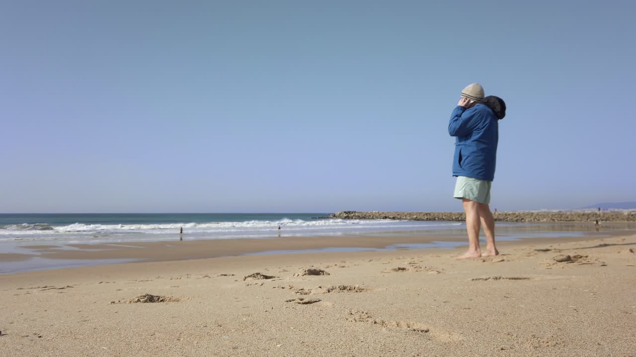 파란색 코트를 입고 해변에서 전화를 받는 나이든 남성