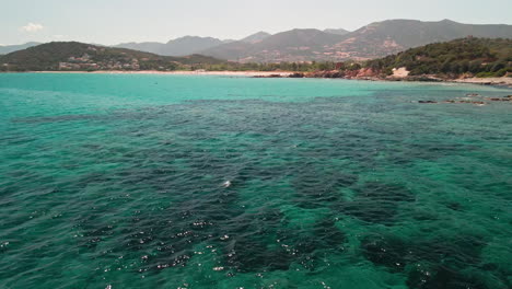 Fliegen-Sie-über-Den-Ruhigen-Tropischen-Ozean-Mit-Bewaldeten-Bergkämmen-Im-Hintergrund-Im-Sommer-In-Sardinien,-Insel-Italien