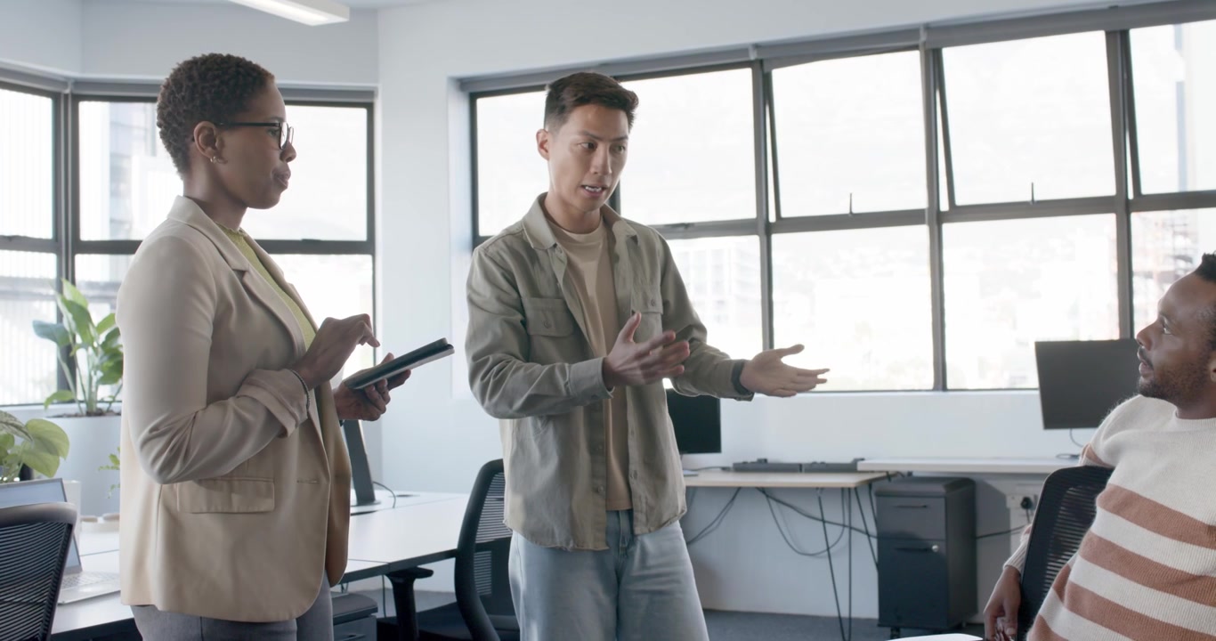 Diverse team reviewing at desk Asian man coming gesturing man typing laptop woman tapping tablet