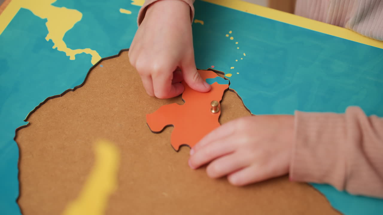 Un estudiante coloca una pieza de puzzle naranja que representa un continente en un mapa, guiando el botón hasta el contorno en agua turquesa, con cuidado para alinearla en la lección de geografía y perfeccionar la motricidad fina.