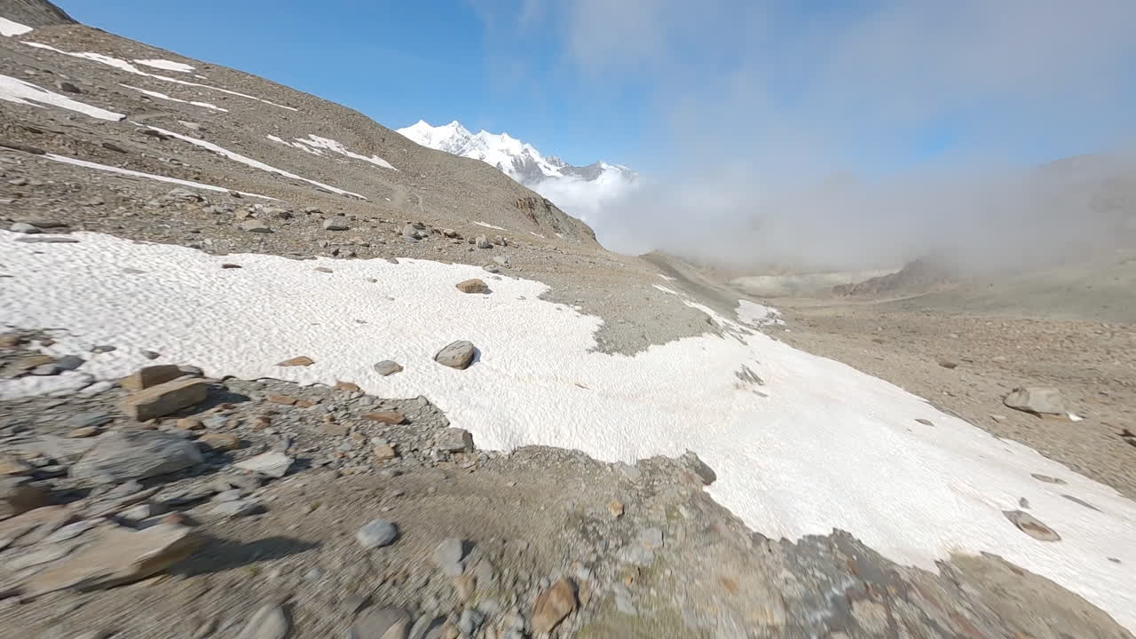 FPV drone follows two mountain bikers descending steep, exposed alpine trails above Saas-Grund and Saas-Fee on a clear bluebird summer day, showcasing epic terrain and speed