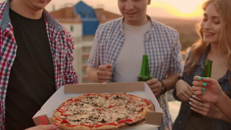 jóvenes chicas y chicos rusos con camisas a cuadros analizan pedazos de pizza caliente con papel rojo y comen juntos. esta es una fiesta en la azotea con una cerveza.