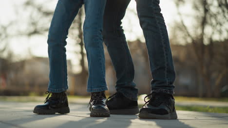 a man is dropping a little girl down. the girl is wearing blue jeans and black boots, and the man's legs are visible in the frame. they are outdoors, with trees and sunlight in the background