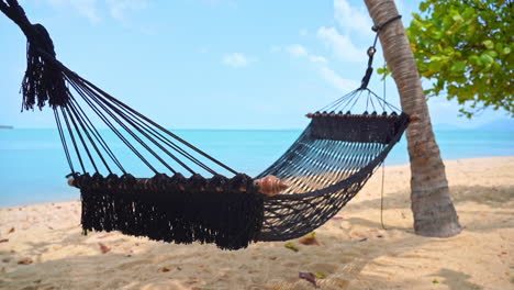 empty cotton rope net hammock with spreader bars lashed between two palm trees on the sandy beach in front of the turquoise sea on a tropical island