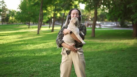 Beautiful-woman-with-dreadlocks-and-headphones-whirling-around-in-the-park-while-holding-her-pet-small-pug
