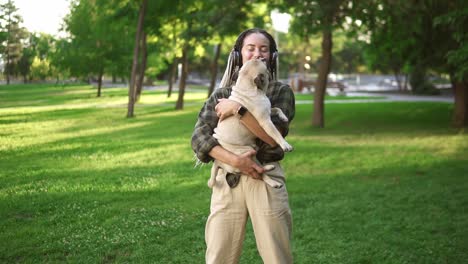 Hermosa-Mujer-Con-Rastas-Y-Auriculares-Dando-Vueltas-En-El-Parque-Mientras-Sostiene-A-Su-Mascota-Pequeño-Pug