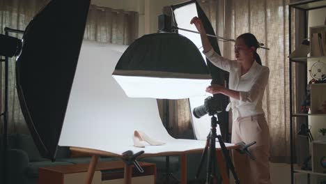 asian female photographer adjusting light equipment to get good illumination on women's shoes while taking photos of the products in home studio