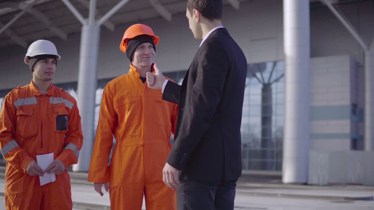 gerente en traje dando sobres con dinero a los trabajadores en uniforme naranja y cascos. finalización exitosa del proyecto. filmado en 4k