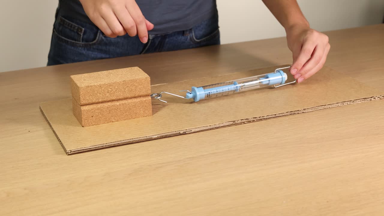 Hands manipulate a wooden block on cardboard using a hook and screw to explore friction dynamics