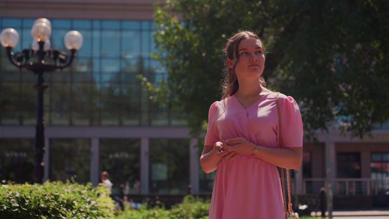 Junge Frau im rosa Kleid steht in einem sonnendurchfluteten Park in der Nähe eines modernen Glasgebäudes, die Hände gefaltet, geflochtene Haare umrahmen das Gesicht, altmodischer Laternenpfahl und üppiges Grün, weiche Schatten und eine sanfte Brise, nachdenklich