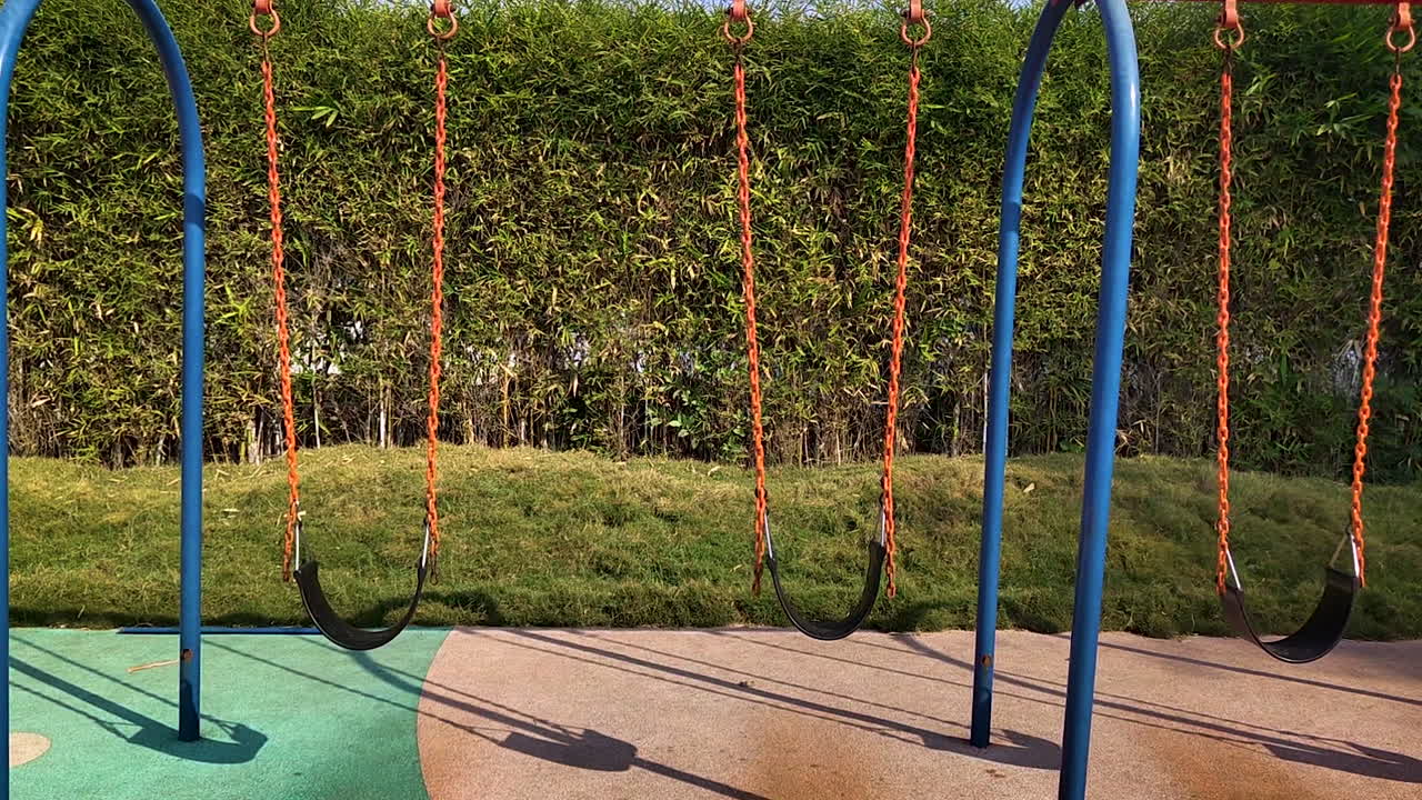 Altalene del parco giochi che si muovono lentamente al vento all'interno di un parco. Sedili delle altalene per bambini vuoti che proiettano ombre sul terreno, con uno sfondo di siepe verde e un'atmosfera calma e soleggiata.