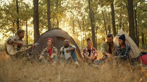 a company of 6 trekking participants in special hiking clothes against the backdrop of tents. a blond man in a hat plays an acoustic guitar and all the other participants in the hike sing along and palm him off against the backdrop of a green, sunny summer forest