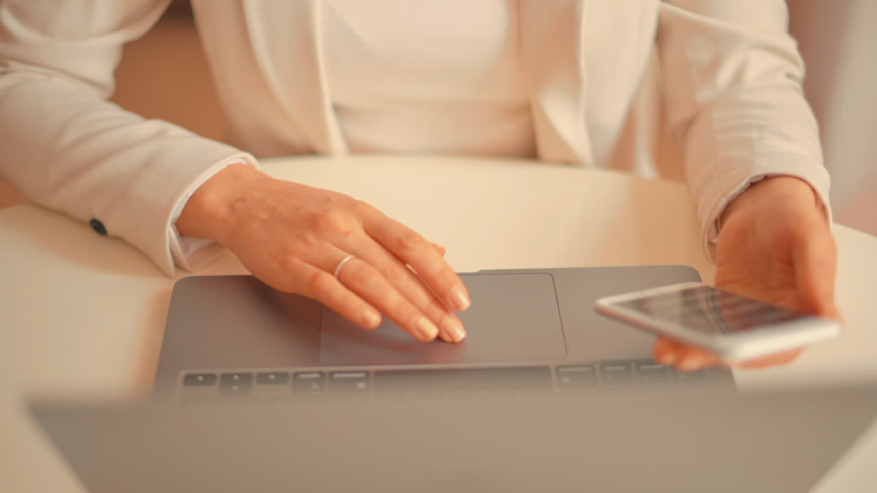 Donna con blazer bianco che lavora a un tavolo su un laptop mentre tiene in mano il telefono
