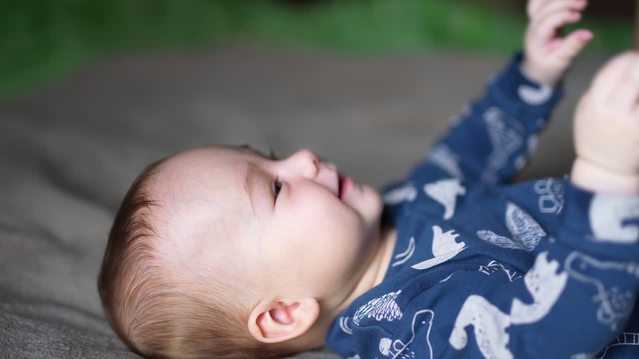 Healthy active toddler boy lies on bed moving and smiling. Cute child stretching hands upwards. Side view. Close up.