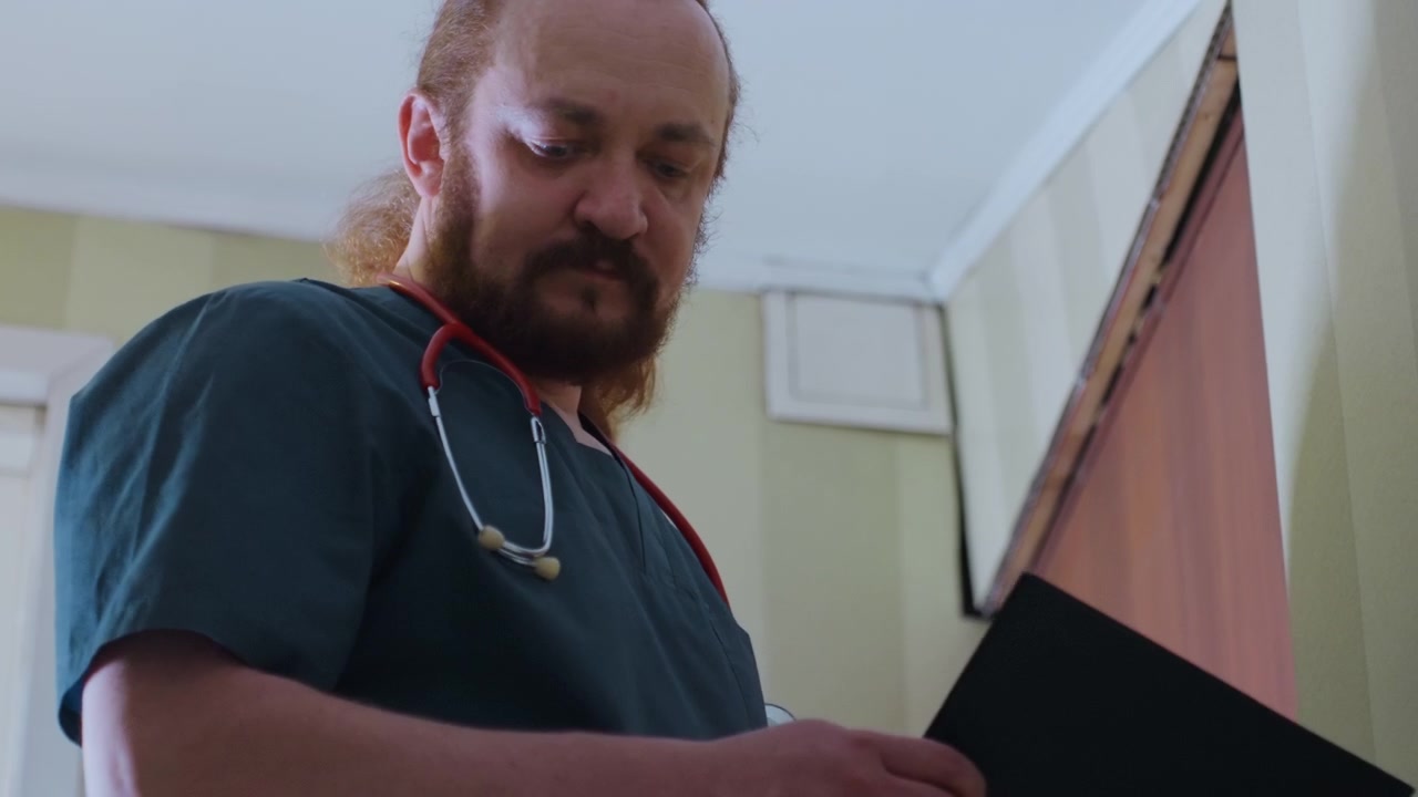 Medico bianco che valuta la cartella del paziente e i parametri vitali, clinico dai capelli rossi in camice verde acqua con stetoscopio che studia una cartella clinica sulla soglia, scena interna concentrata che mostra una valutazione professionale di visita a domicilio.