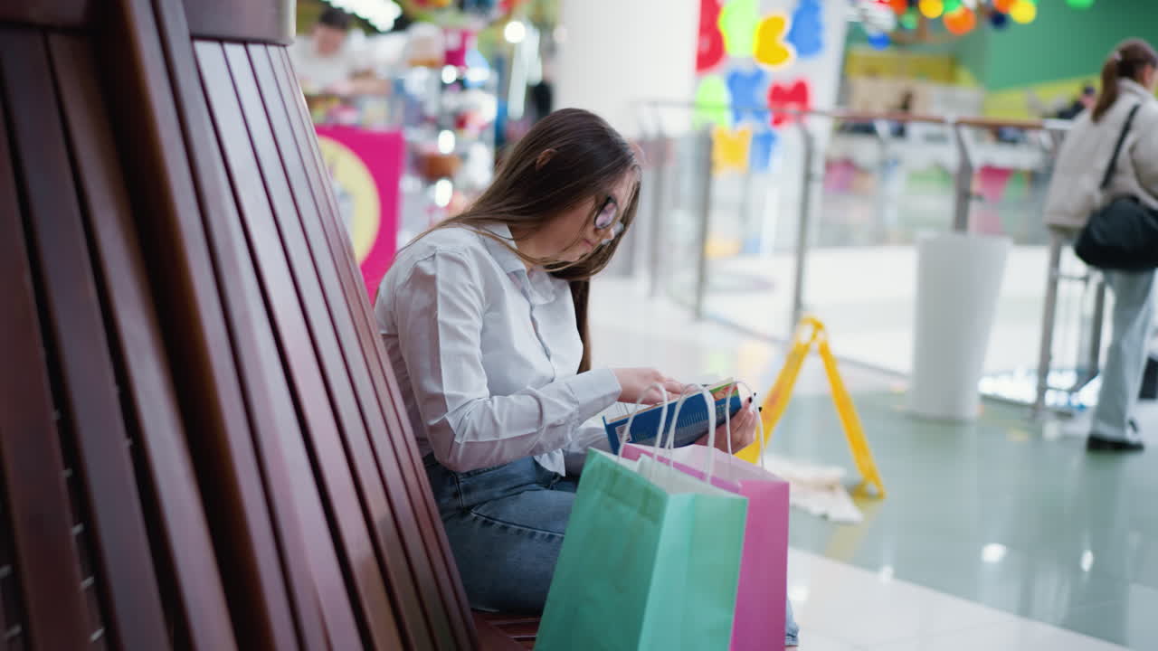 활기차고 바쁜 쇼핑몰의 나무 벤치에 앉아서 독서에 몰두하는 젊은 성인, 에 쇼핑 가방, 흐릿한 배경