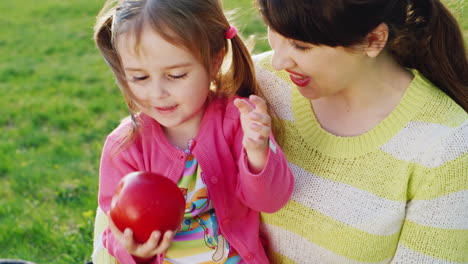 Ein-Kleines-Mädchen-Sitzt-In-Den-Armen-Ihrer-Mutter-Und-Spielt-Mit-Einem-Roten-Reifen-Apfel