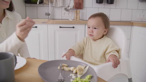 linda niñita sentada en una silla alta en la cocina mientras su madre le da de comer a 2