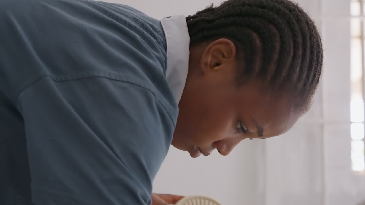 Woman cleaning dishes in house leaning over sink with braided hair and blue uniform, soft sunlight through window, focused expression, washing plate with gentle scrubbing motion, tidy interior, calm