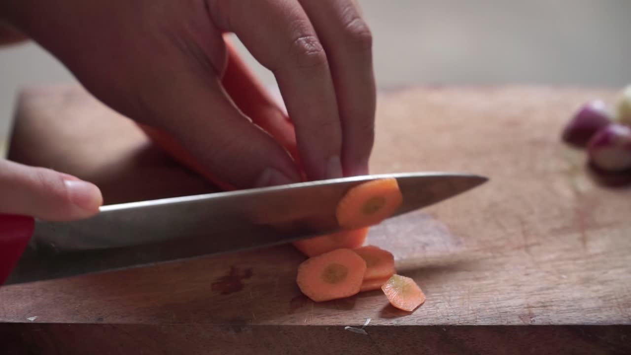 Close-up of hands slicing a fresh carrot on a wooden cutting board. The scene highlights the preparation process in a natural kitchen setting with soft lighting