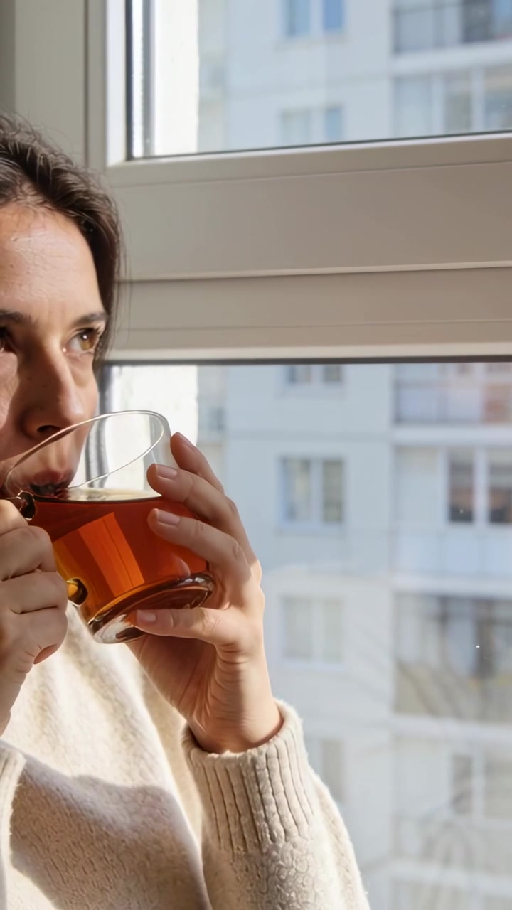 Video verticale: Donna che guarda fuori e sorseggia da una tazza di vetro alla finestra, osservando la facciata con un maglione, spazio per il testo.