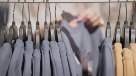 close up of thick polo knit hanging on black hangers in brightly lit store as female shopper carefully selects garment from rack, fingers touching fabric while pulling one polo out to inspect fit