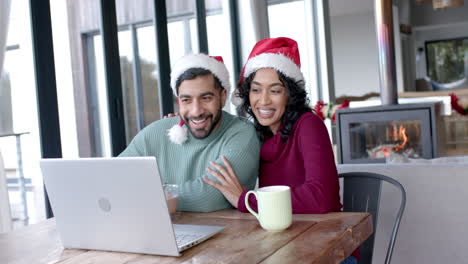 Happy-biracial-couple-wearing-santa-claus-hats-using-laptop-for-video-call-at-home,-in-slow-motion