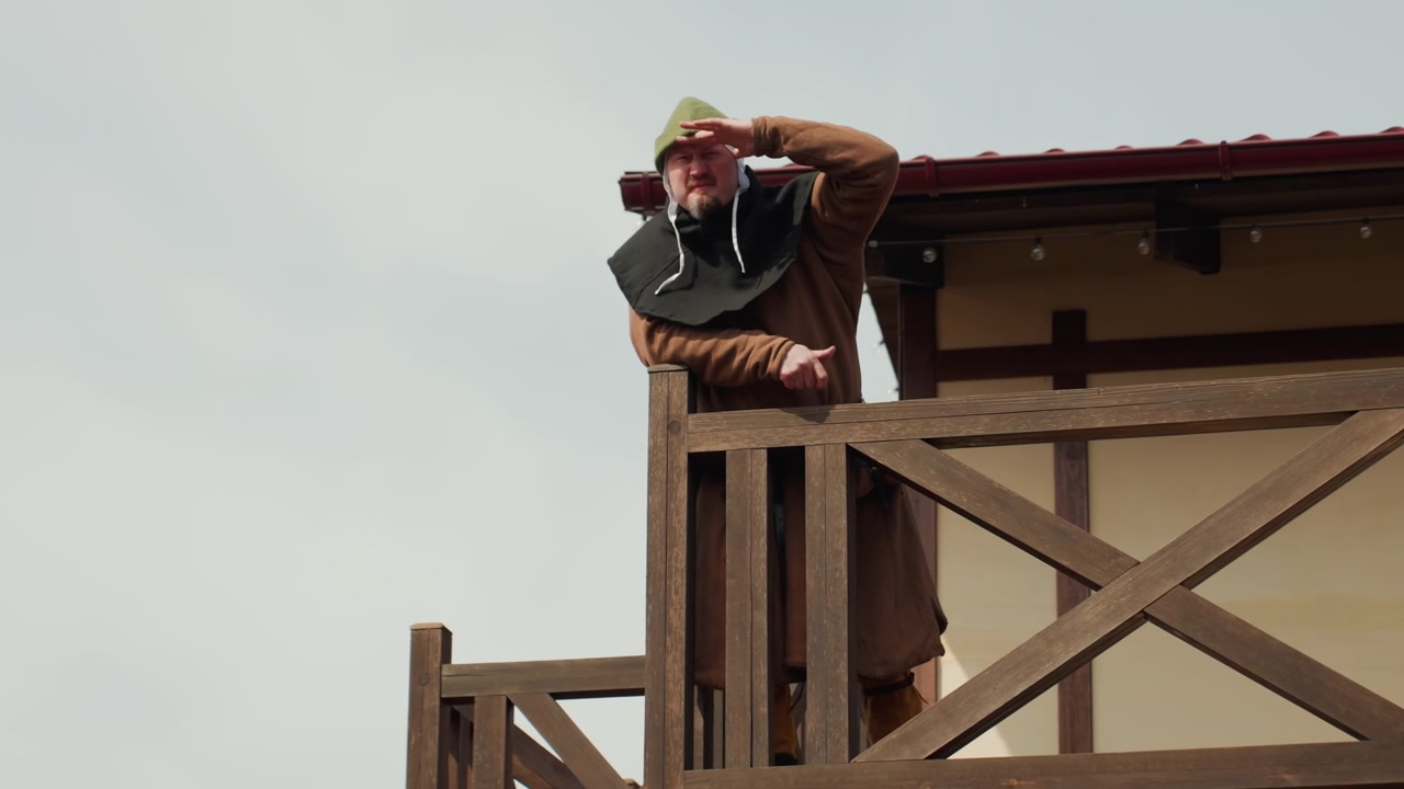 Older man in brown coat and flat cap peers through binoculars while standing on rustic cross braced wooden observation platform, watching distant horizon under soft overcast light beside weathered