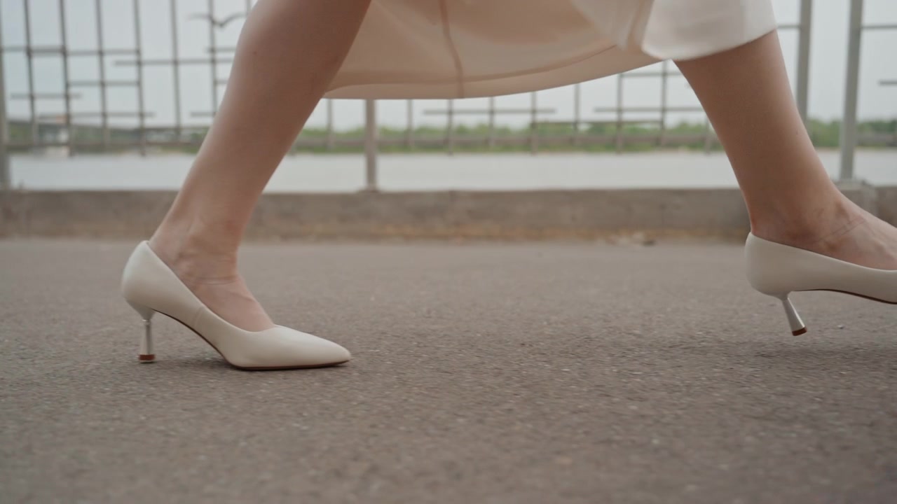 Woman in white dress walks along outdoor path near water with views of her legs and feet as woman moves. background features metal railings and distant greenery under soft daylight