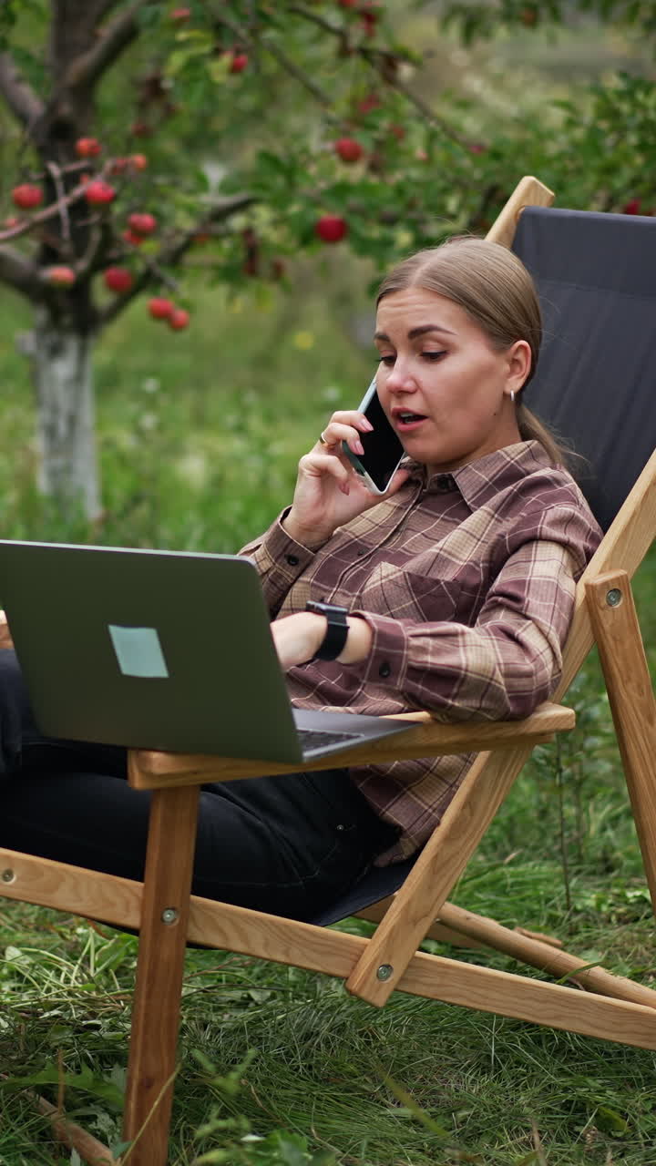 Freelance female specialist sitting in garden chair with laptop and phone. Lady is having conversation over the phone looking at computer screen. Vertical video