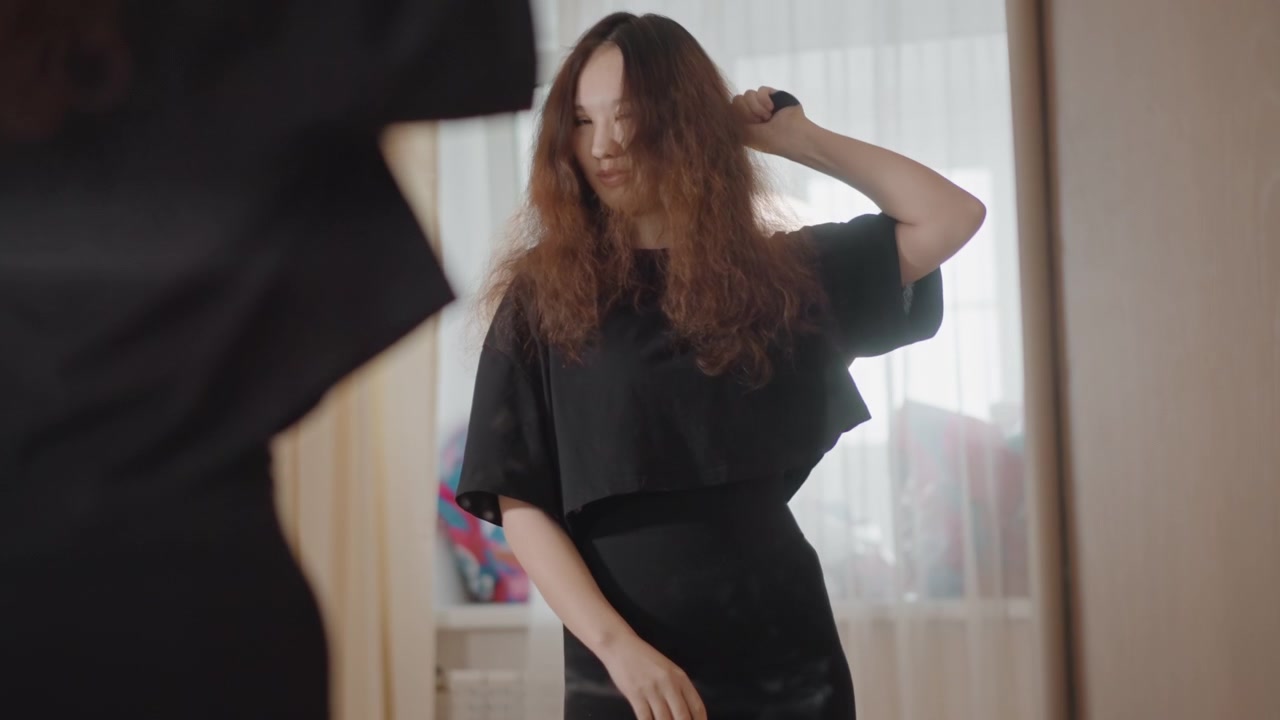 Young woman styles long curly hair before mirror in bright dressing area, using hairdryer and brush while leaning forward with lifted strands, soft window backlight, ring light glow, and relaxed